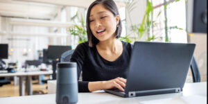 Asian businesswoman talking to virtual assistant at her desk. Female professional working on laptop and talking into a speaker.